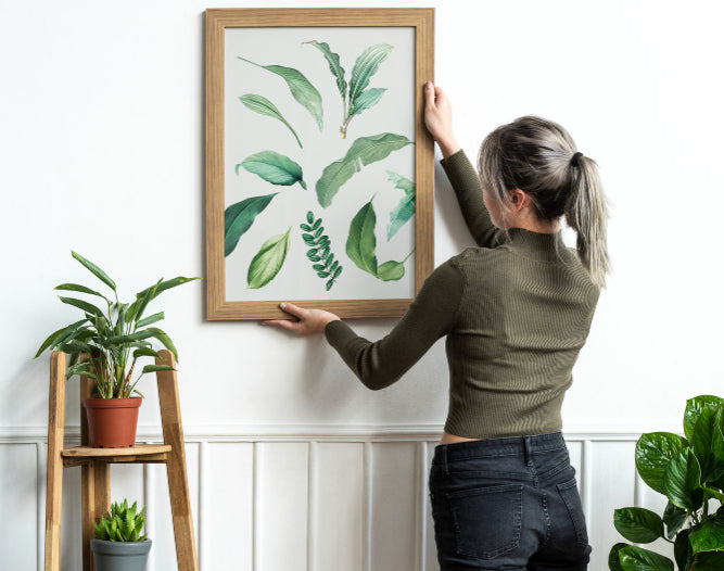 Person hanging a framed artwork of green leaves on a white wall with plants around.